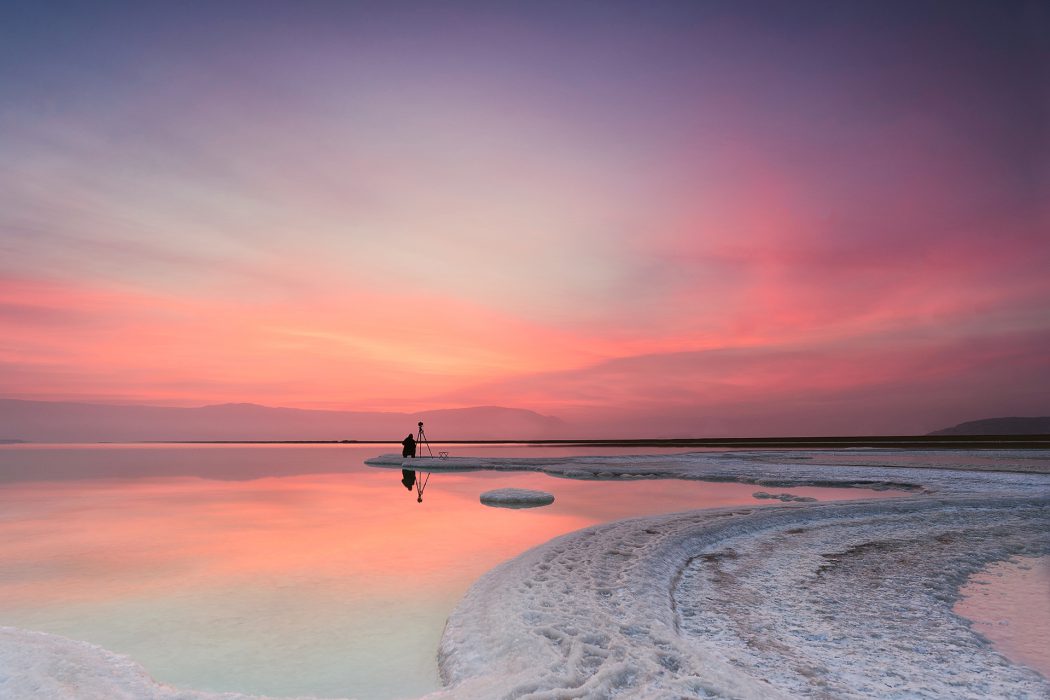A photographer man shooting the landscape on the edge of the salt shore in the Dead Sea at sunrise with beatiful colors
